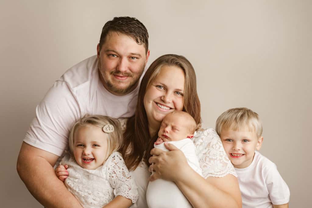 family photo with newborn everyone wearing white. Dads arms around mom with little girl, little boy and newborn baby boy