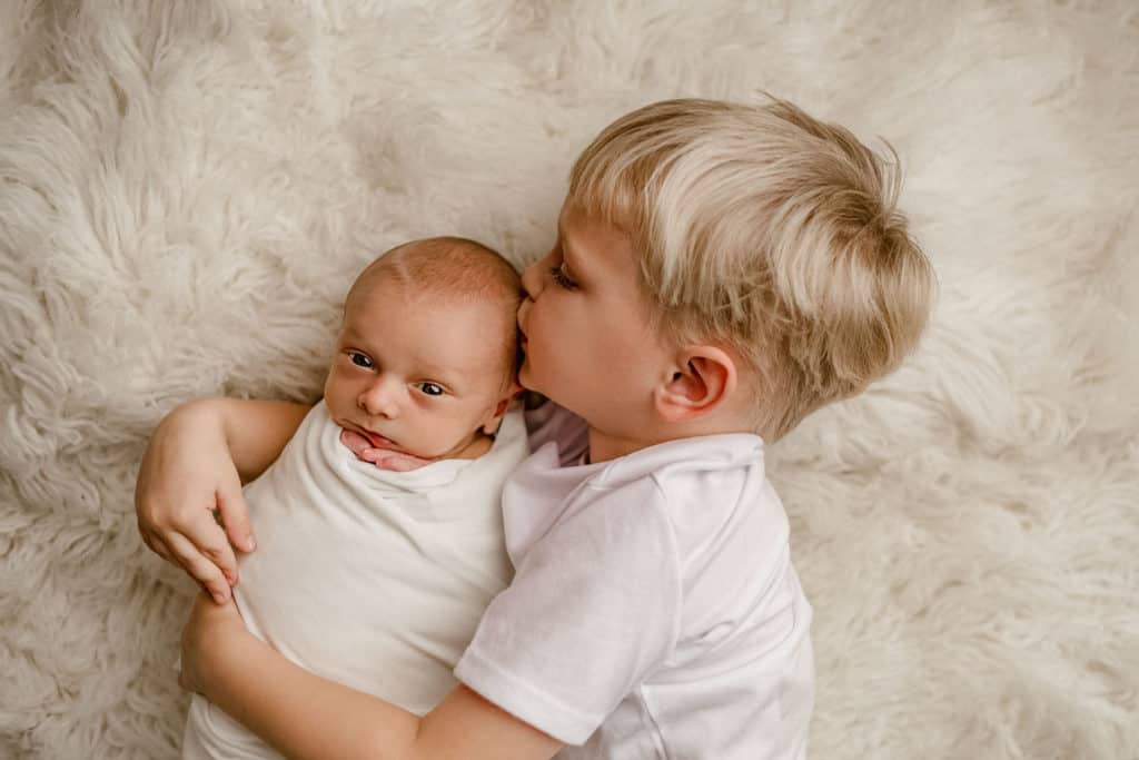 big brother giving newborn baby brother kiss on white fur