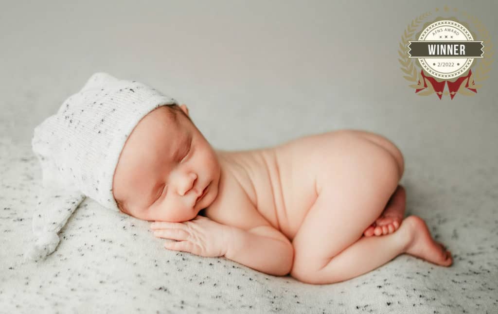 newborn baby boy posed for photo on cream speckled background wearing sleepy hat