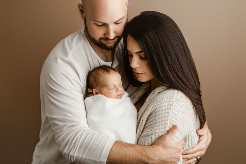 family posed for newborn photo on tan background. mom and dad holding newborn baby and smiling down at him bundled in white